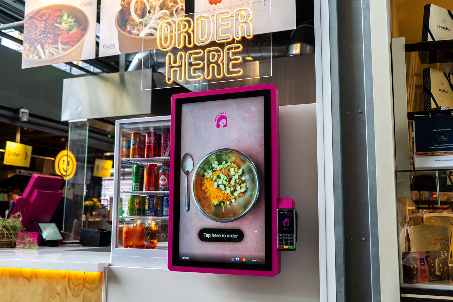 Self-service digital ordering screen with a bowl of salad and a spoon, next to a drink refrigerator and a neon sign reading 'ORDER HERE'.