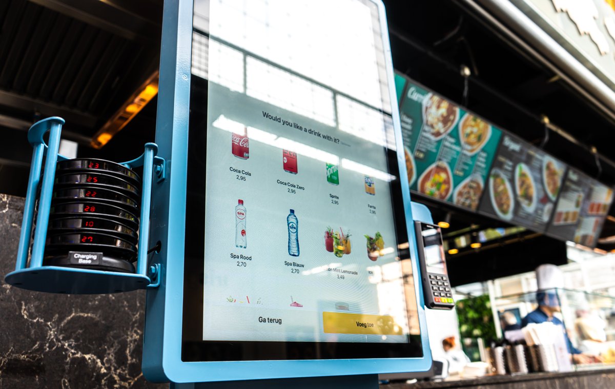 Onesix Kiosk Premium dans une halle gourmande animée, avec une question d'upsell en anglais sur l'écran de commande et des buzzers sur la station de charge