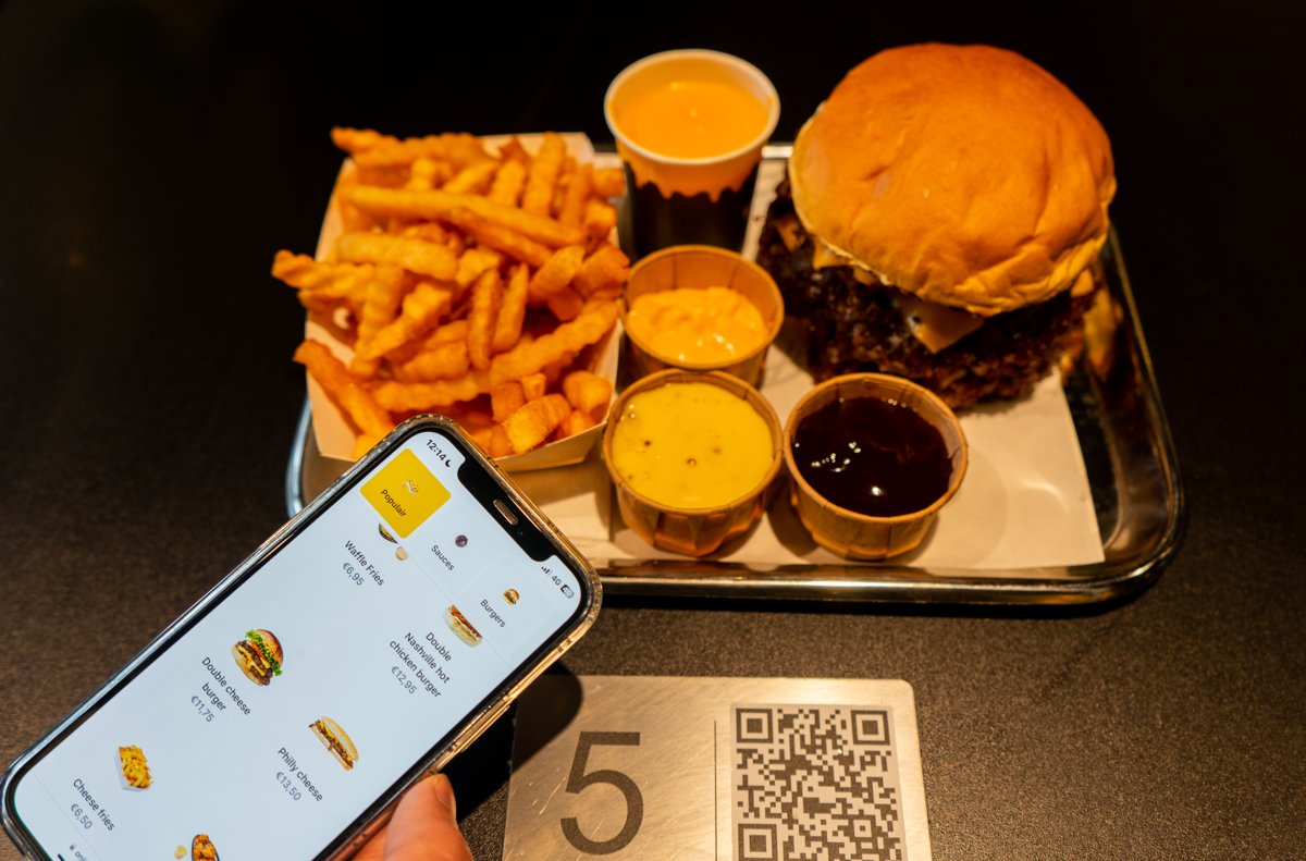 Client consultant le menu digital sur son téléphone, à côté d'une plaque de table avec numéro et QR code, avec un burger et des frites sur un plateau