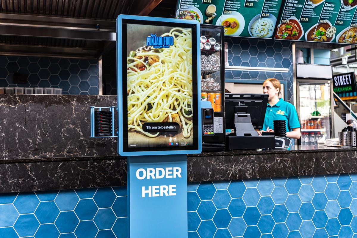 Borne Onesix bleue avec « Order here » à côté du comptoir avec caisse dans un restaurant indien à emporter